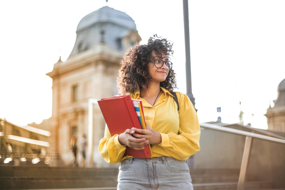 A person with curly hair, wearing glasses and a yellow shirt, stands outdoors holding red folders, with a historic building in the background.